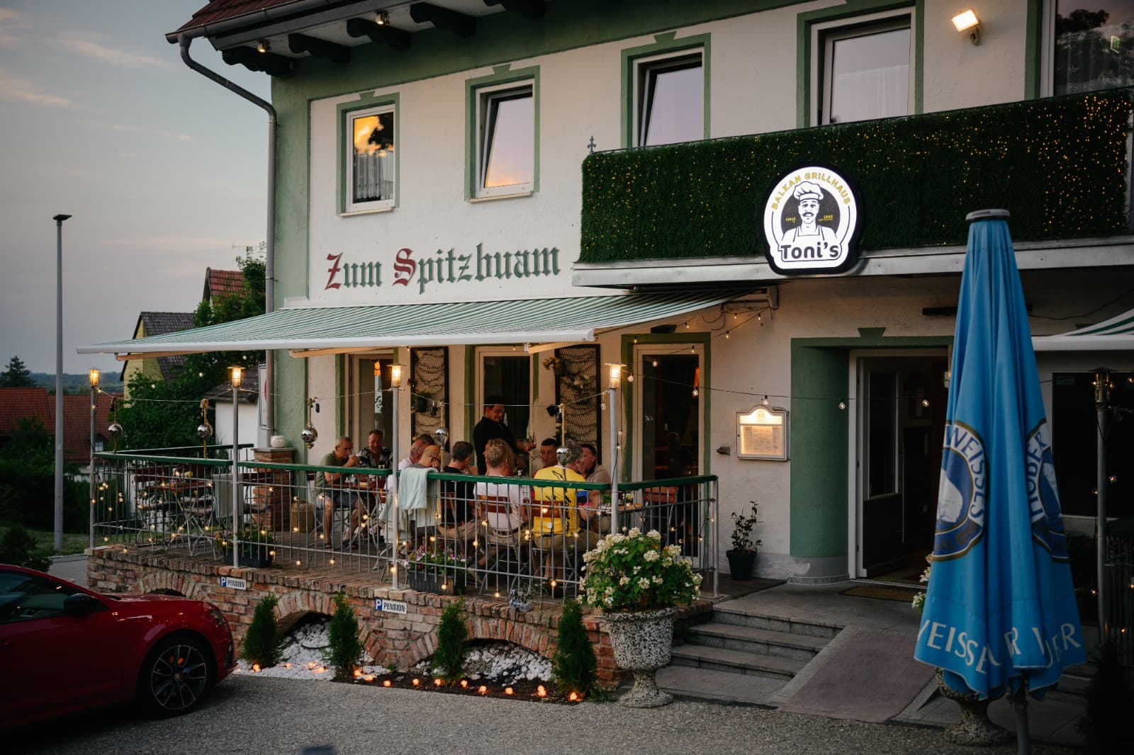 Toni's Balkan Grillhaus in Attenkirchen bei Dämmerung, Terrasse mit Gästen und warmem Lichterglanz
